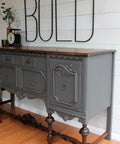 Dixie Belle Paint Gravel Road Chalk Mineral Paint is the perfect paint for any DIY project! Credenza painted in Gravel Road Chalk Mineral Paint with dark brown wood on top and on the legs of the piece with bronze hardware to enhance the piece. Staged with an antique food scale, bronze cup and copper vase with leaf branches inside. Large black letters above piece say "BOLD".