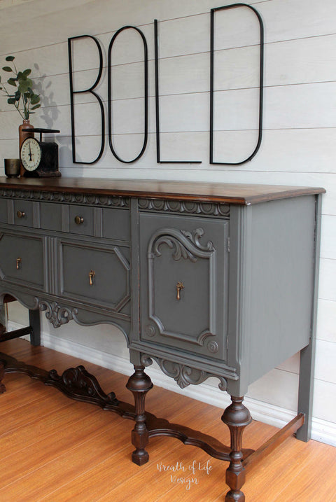 Dixie Belle Paint Gravel Road Chalk Mineral Paint is the perfect paint for any DIY project! Credenza painted in Gravel Road Chalk Mineral Paint with dark brown wood on top and on the legs of the piece with bronze hardware to enhance the piece. Staged with an antique food scale, bronze cup and copper vase with leaf branches inside. Large black letters above piece say "BOLD".