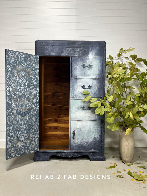 Dixie Belle Paint Gravel Road Chalk Mineral Paint is the perfect paint for any DIY project! a Wardrobe with door open painted in Gravel Road Chalk Mineral Paint with distressed white blue paint on drawers that are on the right of the door. A distressed Belles & Whistles Stencil in Morocco is seen on the inside of door. Staged next to a brown vase with large leaves on branches on light brown floor with white wall.