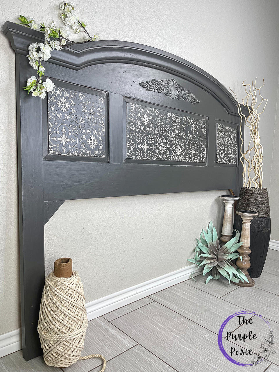 Dixie Belle Paint Gravel Road Chalk Mineral Paint is the perfect paint for any DIY project! Headboard painted in Gravel Road Chalk Mineral Paint with Belles & Whistles Sonoma Art Tile - Stencil along the center in white. Staged against a white wall with large roll of twine on left side and two large candle holders with a large brown vase with white branches inside beside the candle holders. A branch of flowers hang above furniture.