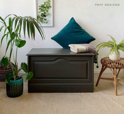 Dixie Belle Paint Gravel Road Chalk Mineral Paint is the perfect paint for any DIY project! Chest painted in Gravel Road Chalk Mineral Paint staged with blue pillow and blanket on top right side, On top of brown rug with houseplants on each side of piece.