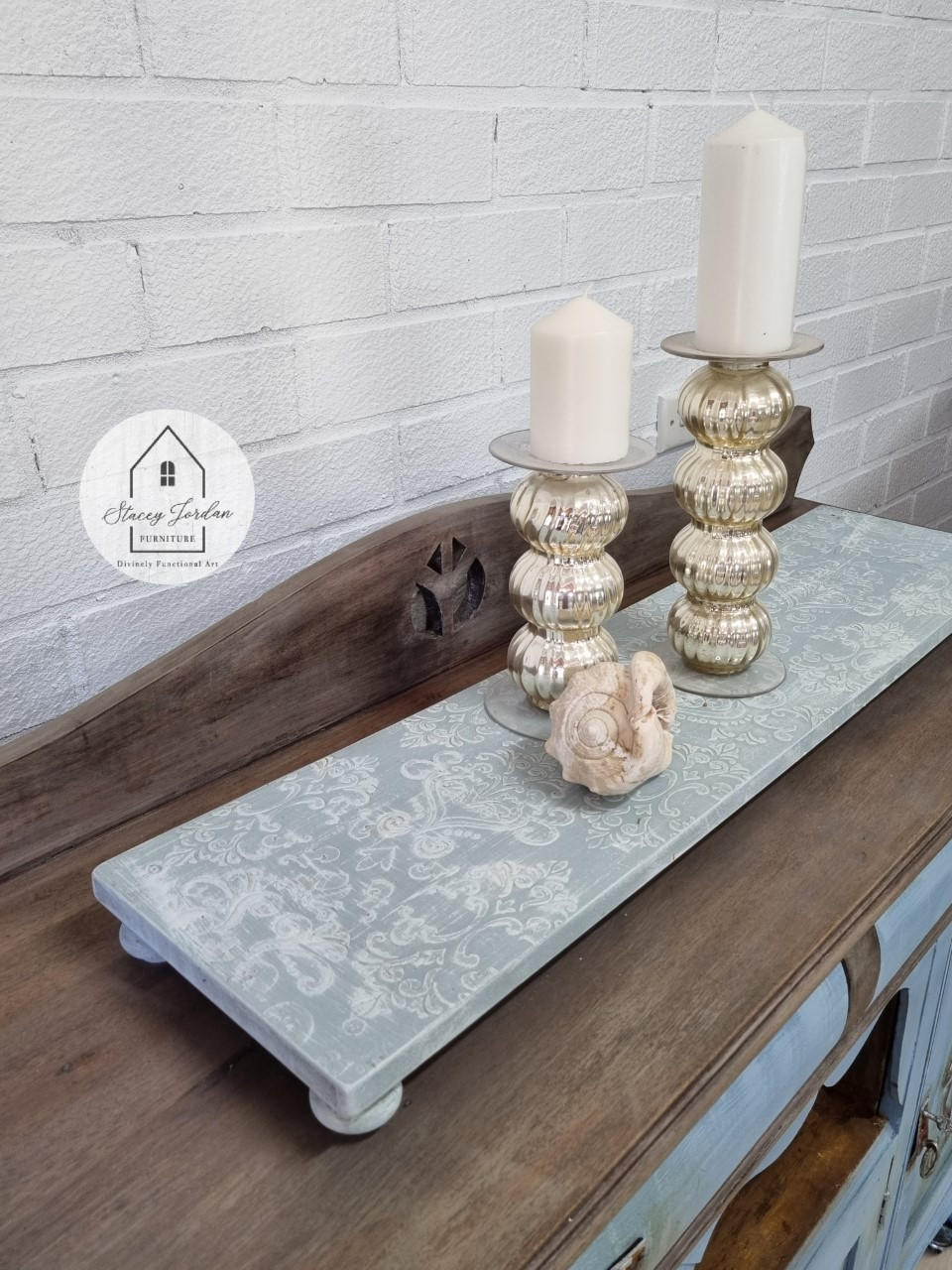 Dixie Belle Paint Haint Blue Chalk Mineral Paint is the perfect paint for any DIY project!
A wooden tray with legs painted with Blue Chalk Mineral Paint  and the Victorian Damask - Stencil in white is seen on the top side of the tray. Staged with two gold candle pedestals with white candles along with a seashell.