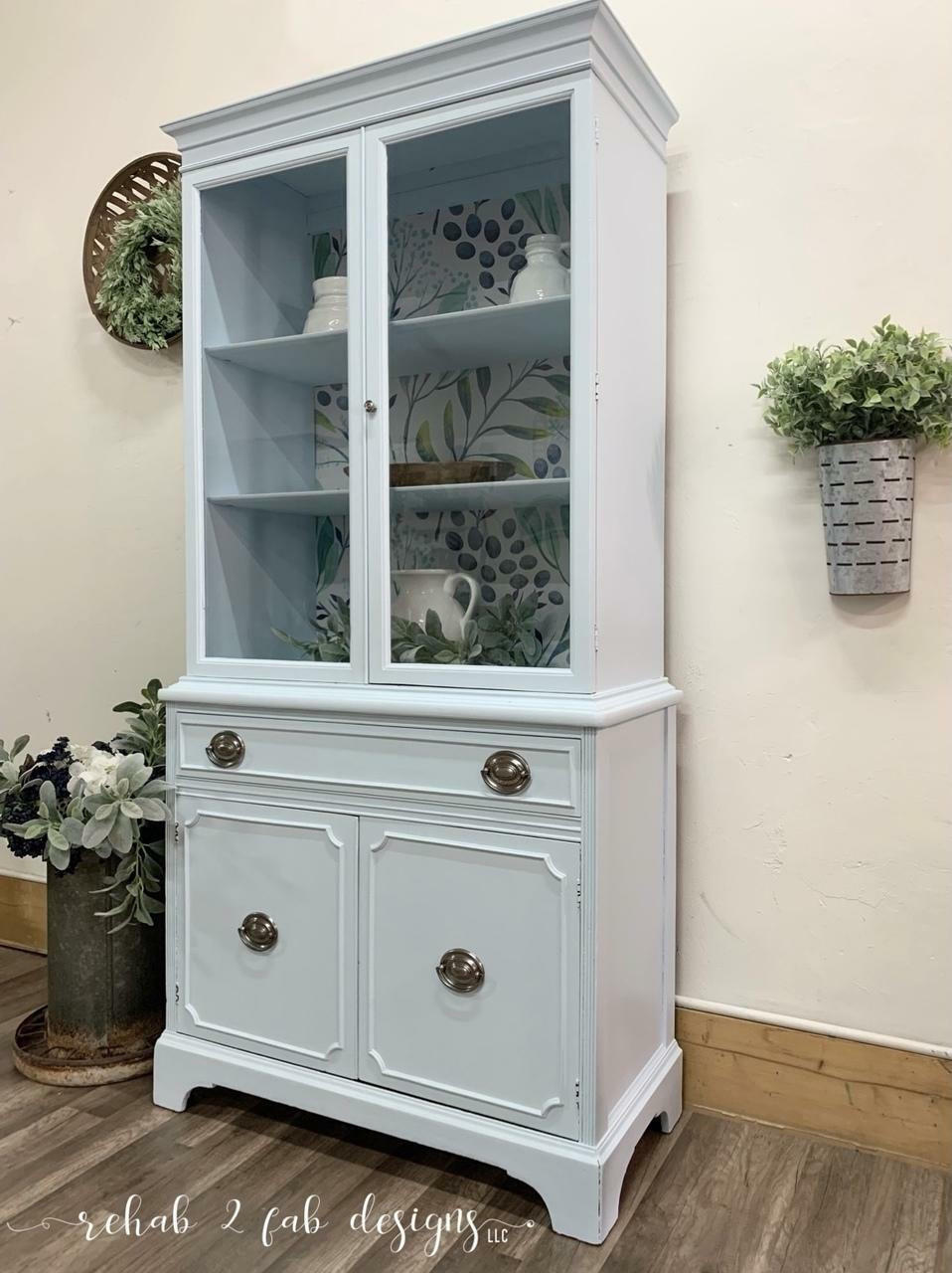 Dixie Belle Paint Haint Blue Chalk Mineral Paint is the perfect paint for any DIY project!
A kitchen hutch painted in Blue Chalk Mineral Paint with silver hardware. Stage with pitchers and teapots inside the hutch, As well as greenery around the piece.