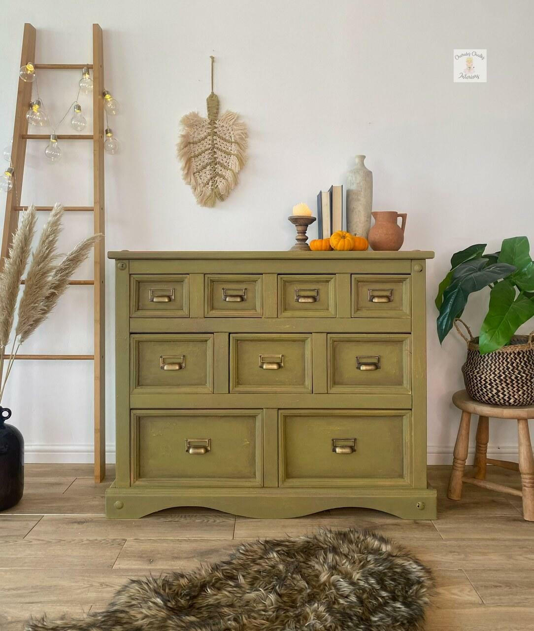 Dixie Belle Paint Holy Guacamole Chalk Mineral Paint is the perfect paint for any DIY project!
Dresser painted with Holy Guacamole Chalk Mineral Paint with bronze hardware staged with feathers, greenery around the piece, a staged latter with lights hanging off the ladder. On top of the furniture are books, vase and miniature 3 pumpkin. A fur gray rug is seen in front of the piece.
