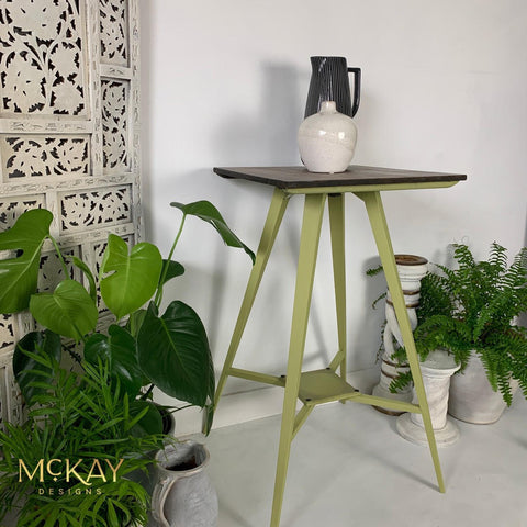 Dixie Belle Paint Holy Guacamole Chalk Mineral Paint is the perfect paint for any DIY project!
A table painted in Holy Guacamole Chalk Mineral Paint with a dark brown top. Staged with potted house plants around the piece with a black pitcher and a white vase on top of the table. A white wall and white floor room.