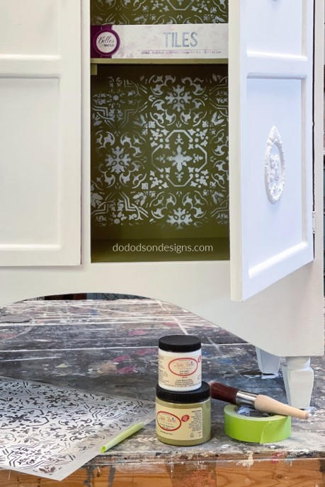 Dixie Belle Paint Holy Guacamole Chalk Mineral Paint is the perfect paint for any DIY project!
A white opened cabinet with the inside of the cabinet painted in Holy Guacamole Chalk Mineral Paint with the Sonoma Art Tile - Stencil in white all over the inside. Staged with Dixie Belle Paint Company product that were used on the piece which were, Cotton CMP, Holy Guacamole Chalk Mineral Paint, DBP Synthetic Brush, and the Moroccan -Stencil, all in front and on the floor of the piece.