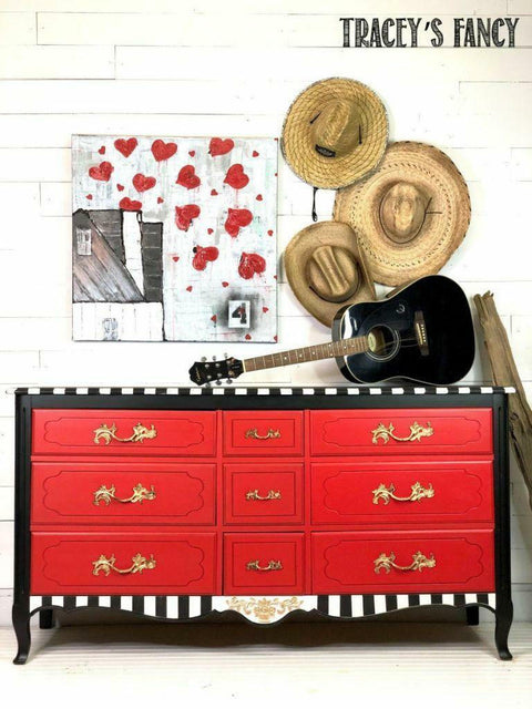 Dixie Belle Paint Honky Tonk Red Chalk Mineral Paint is the perfect paint for any DIY project!
Dresser painted with the drawers painted in Honky Tonk Red Chalk Mineral Paint and the body painted in black with the trims as white & black stripes. Staged with a black acoustic guitar and three straw hats on the wall hanging above the piece to the left has a painting with red hearts.