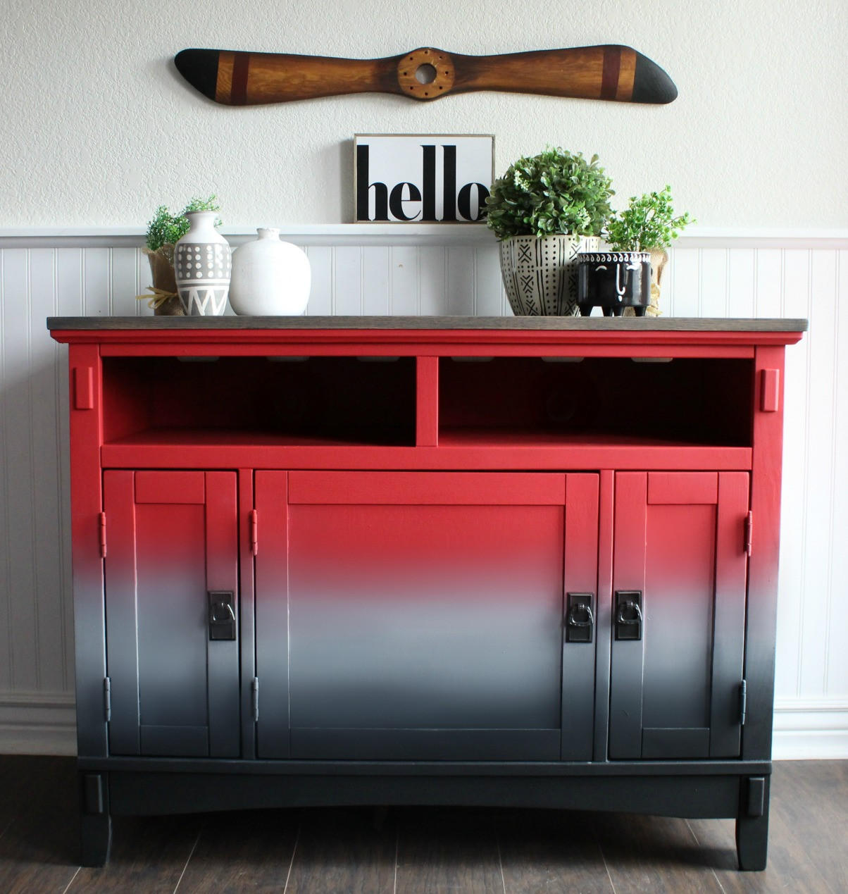 Dixie Belle Paint Honky Tonk Red Chalk Mineral Paint is the perfect paint for any DIY project!
A credenza with gradient effect with the starting color Honky Tonk Red Chalk Mineral Paint fading into gray then black with black hardware. Staged on top of the piece are potted houseplants and vases with a sign above the piece that reads "HELLO".