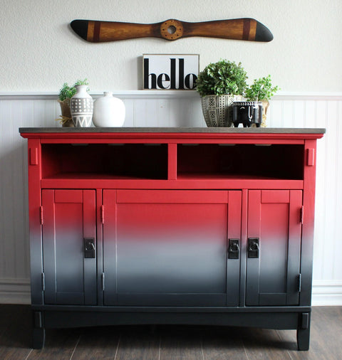 Dixie Belle Paint Honky Tonk Red Chalk Mineral Paint is the perfect paint for any DIY project!
A credenza with gradient effect with the starting color Honky Tonk Red Chalk Mineral Paint fading into gray then black with black hardware. Staged on top of the piece are potted houseplants and vases with a sign above the piece that reads "HELLO".