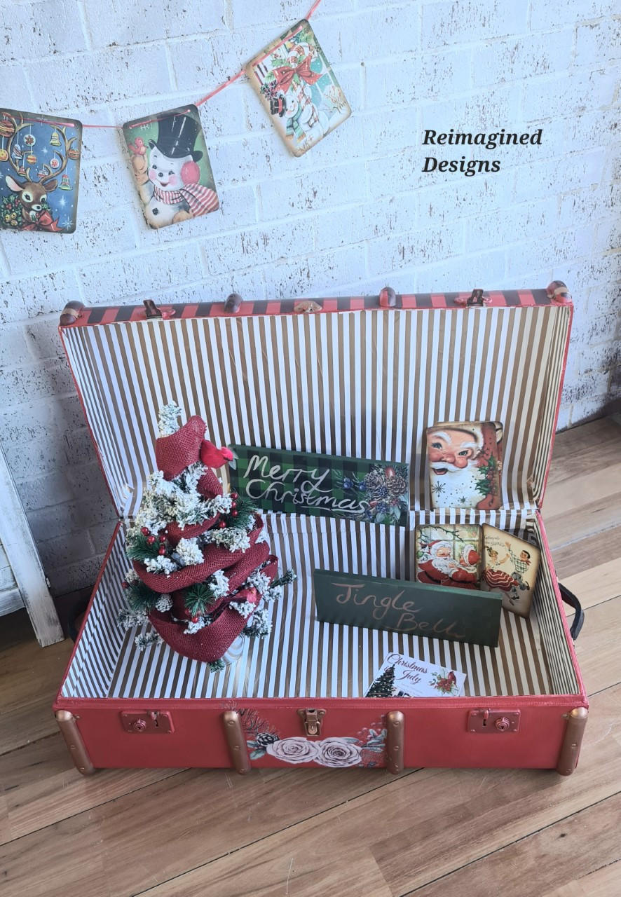 Dixie Belle Paint Honky Tonk Red Chalk Mineral Paint is the perfect paint for any DIY project!
An opened vintage luggage painted in Honky Tonk Red Chalk Mineral Paint with gold accents and inside the luggage are white and gold stripes staged with Christmas niknaks  like small signs that read "MERRY CHRISTMAS" " JINGLE BELL".