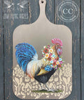 Dixie Belle Paint Hurricane Gray Chalk Mineral Paint is the perfect paint for any DIY project!
A wooden brown serving board with hand painted rooster and a flower necklace on the background of the rooster is the Tea Towel Floral - Stencil in a light tan. Staged hanging on a black distressed wood wall.