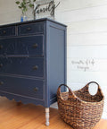Dixie Belle Paint In the Navy Chalk Mineral Paint is the perfect paint for any DIY project!
A Dresser painted in In the Navy Chalk Mineral Paint  with distressed edges around the dresser. Staged with a woven basket besides the piece along a white wall.