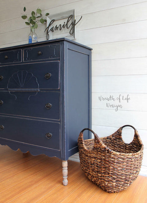 Dixie Belle Paint In the Navy Chalk Mineral Paint is the perfect paint for any DIY project!
A Dresser painted in In the Navy Chalk Mineral Paint  with distressed edges around the dresser. Staged with a woven basket besides the piece along a white wall.