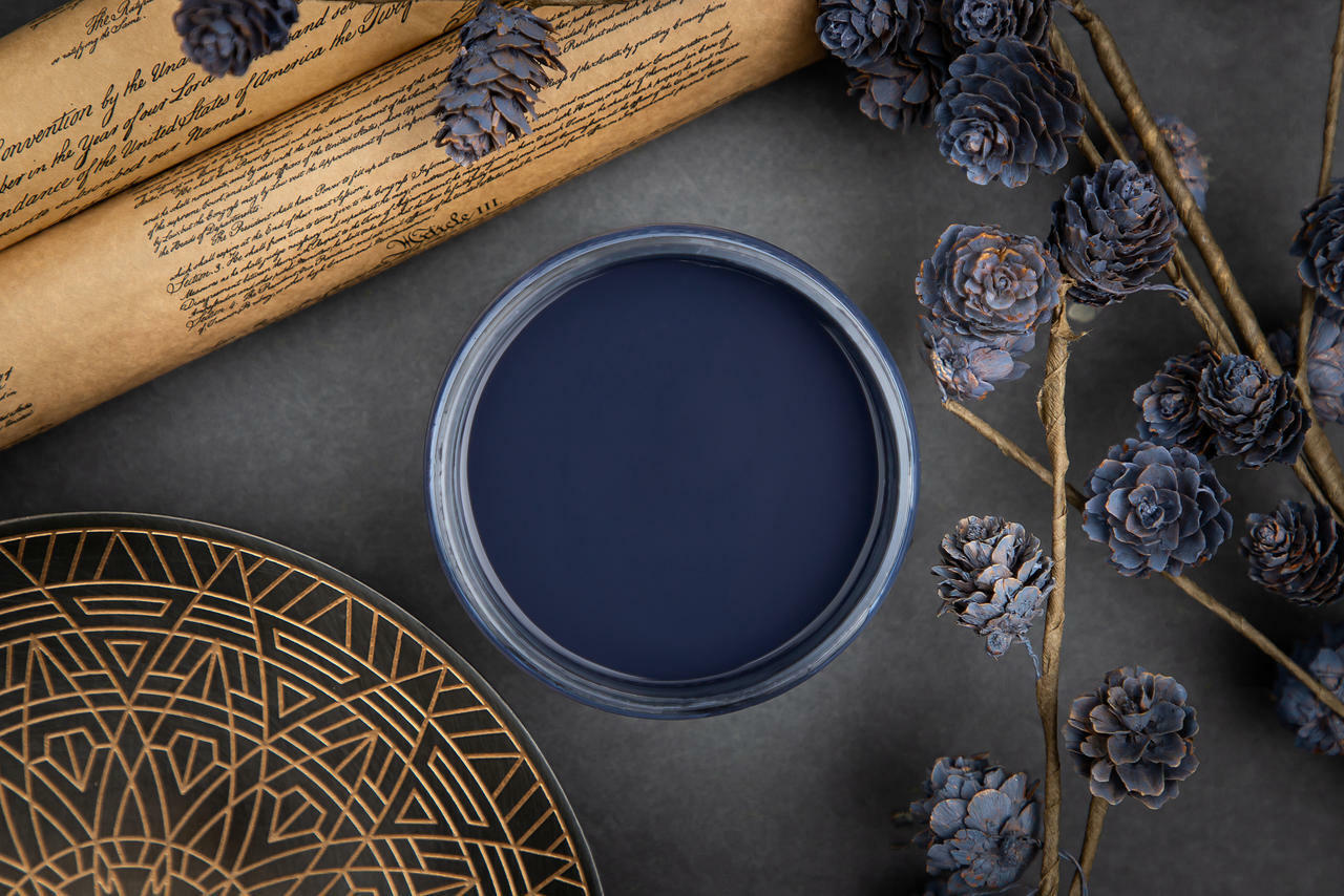 Dixie Belle Paint In the Navy Chalk Mineral Paint is the perfect paint for any DIY project!
A top view of an opened container of In the Navy Chalk Mineral Paint, staged with pinecones painted with In the Navy CMP along with scrolls and a black and old plate.