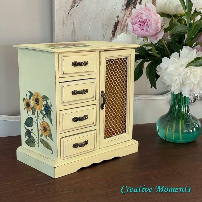 Dixie Belle Paint Lemonade Chalk Mineral Paint is the perfect paint for any DIY project!
Jewelry box painted with black hardware and the Sunflower Transfer is seen on the top of the piece and on the side panel as well. Staged besides a glass vase of white and pink flowers on a brown wood platform.