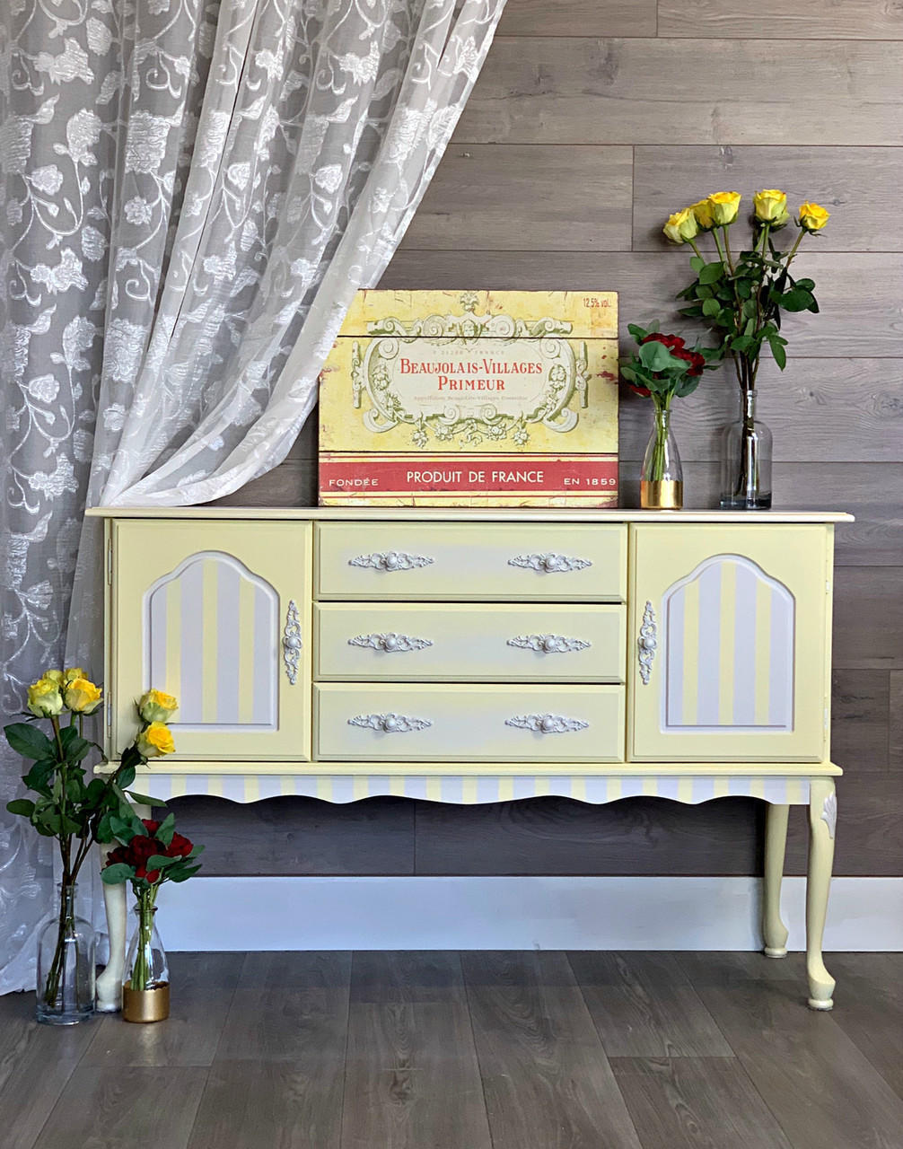 Dixie Belle Paint Lemonade Chalk Mineral Paint is the perfect paint for any DIY project!
Credenza painted in Lemonade Chalk Mineral Paint with gray stripes on the door panels and trim. along with white hardware, staged with glass bottles and yellow/red roses inside them.