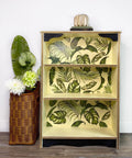 Dixie Belle Paint Lemonade Chalk Mineral Paint is the perfect paint for any DIY project!
Bookshelf case painted in Lemonade Chalk Mineral Paint with green leaves of tropical plants on the back panel, Staged next to a woven basket with flowers and greenery.