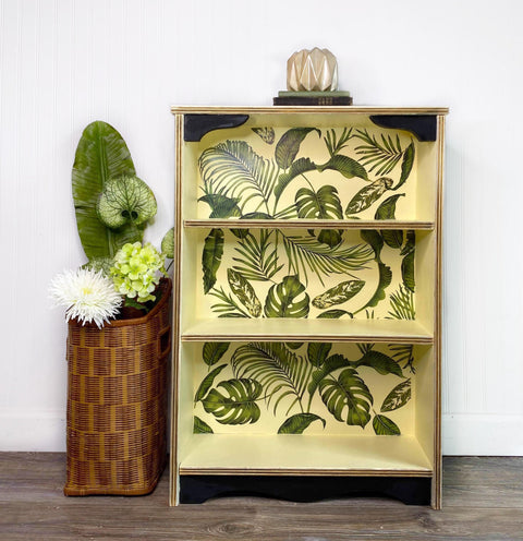 Dixie Belle Paint Lemonade Chalk Mineral Paint is the perfect paint for any DIY project!
Bookshelf case painted in Lemonade Chalk Mineral Paint with green leaves of tropical plants on the back panel, Staged next to a woven basket with flowers and greenery.