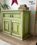 Dixie Belle Paint Limeade Chalk Mineral Paint is the perfect paint for any DIY project!
A Cabinet painted with a mix of Limeade and Evergreen Chalk Mineral Paint, there are off white areas around the edges of the door and the drawer. Staged with a stack of books on top, as well as a ladder with multiple colors hanging on it to the right side of the piece.