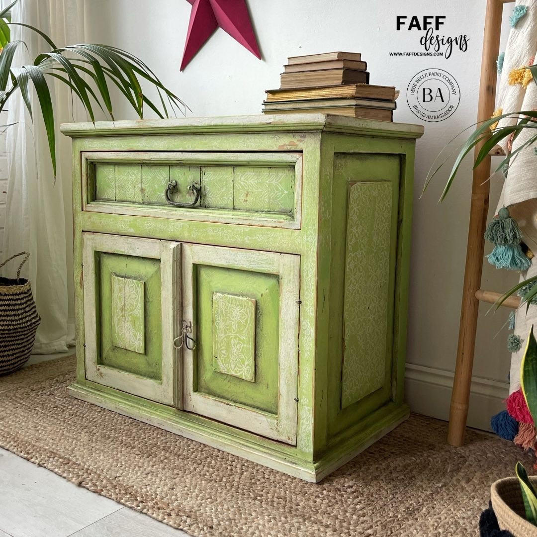 Dixie Belle Paint Limeade Chalk Mineral Paint is the perfect paint for any DIY project!
A Cabinet painted with a mix of Limeade and Evergreen Chalk Mineral Paint, there are off white areas around the edges of the door and the drawer. Staged with a stack of books on top, as well as a ladder with multiple colors hanging on it to the right side of the piece.