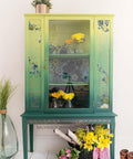 Dixie Belle Paint Limeade Chalk Mineral Paint is the perfect paint for any DIY project!
China Cabinet painted with from top to bottom in a blended fashion,  Limeade, Kudzu and lastly Palmetto Green Chalk Mineral Paint. Staged with small vases with yellow flowers inside piece. As well as a black woven basket with sticks, yellow and pink flowers with a pruning shears and a pair of working boots to the side of the piece.