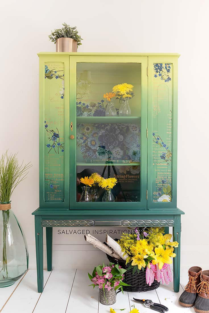 Dixie Belle Paint Limeade Chalk Mineral Paint is the perfect paint for any DIY project!
China Cabinet painted with from top to bottom in a blended fashion,  Limeade, Kudzu and lastly Palmetto Green Chalk Mineral Paint. Staged with small vases with yellow flowers inside piece. As well as a black woven basket with sticks, yellow and pink flowers with a pruning shears and a pair of working boots to the side of the piece.