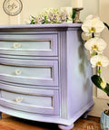Dixie Belle Paint Lucky Lavender Chalk Mineral Paint is the perfect paint for any DIY project!
Short dresser painted in Lucky Lavender Chalk Mineral Paint  with hints of baby blue in front of drawers and around piece. Staged with white flowers around the piece.