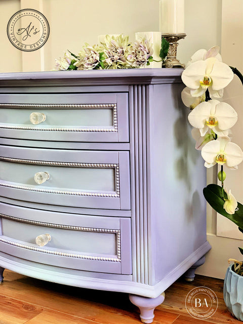 Dixie Belle Paint Lucky Lavender Chalk Mineral Paint is the perfect paint for any DIY project!
Short dresser painted in Lucky Lavender Chalk Mineral Paint  with hints of baby blue in front of drawers and around piece. Staged with white flowers around the piece.