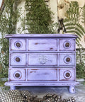 Dixie Belle Paint Lucky Lavender Chalk Mineral Paint is the perfect paint for any DIY project!
Distressed dresser painted in Lucky Lavender CMP with bronze hardware. Staged with greenery around piece.