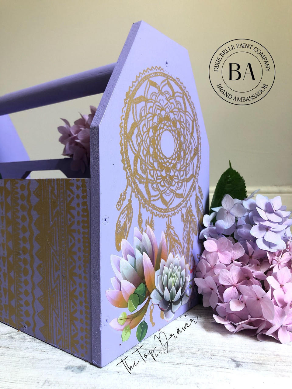 Dixie Belle Paint Lucky Lavender Chalk Mineral Paint is the perfect paint for any DIY project!
Wooden toolbox painted in Lucky Lavender Chalk Mineral Paint with the Mudcloth Stencil on the front of the box and Western Boho - Silkscreen Stencil on the side. Staged with pink & purple flowers.