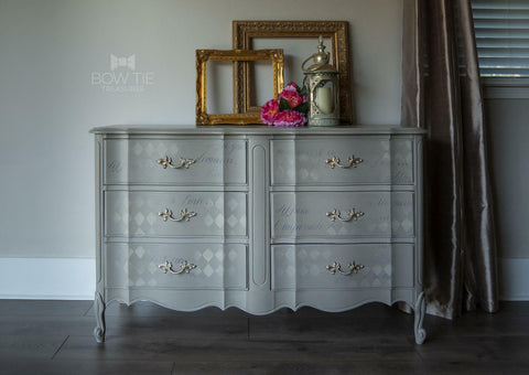 Dixie Belle Paint Mason Dixon Gray Chalk Mineral Paint is the perfect paint for any DIY project! Light Gray dresser chalk painted with Stenciled squares. Staged with gold picture frames, a candle holder with candle and some pink flowers.