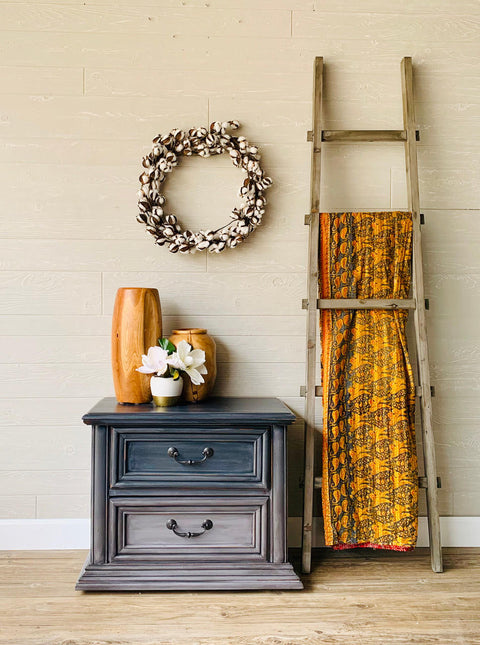 Dixie Belle Paint Mason Dixon Gray Chalk Mineral Paint is the perfect paint for any DIY project! Light gray and dark Blue Chalk painted Ombre end table. Staged with a potted vase with flowers, and some wood vases on top. A cotton wreath is hanging above it and a blanket ladder with a yellow blanket beside it.