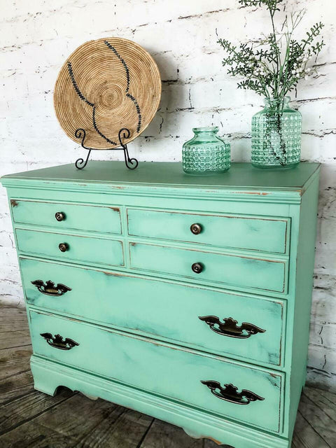 Dixie Belle Paint Mint Julep Chalk Mineral Paint is the perfect paint for any DIY project!
Mint distressed chalk painted dresser with gold hardware. Staged with two glass vases with plants inside and a wicker basket in a plate holder.