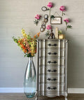 Steel Magnolia Moonshine metallic is the perfect paint to add shimmer and shine to your chalk painted furniture. Grayish metallic chalk mineral painted tall dresser with black hardware. Staged with flowers, tall glass vase, buddha statue and a crystal.