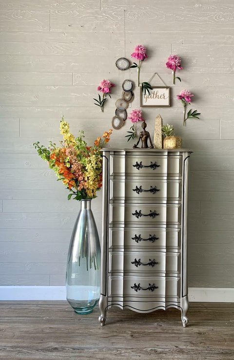 Steel Magnolia Moonshine metallic is the perfect paint to add shimmer and shine to your chalk painted furniture. Grayish metallic chalk mineral painted tall dresser with black hardware. Staged with flowers, tall glass vase, buddha statue and a crystal.
