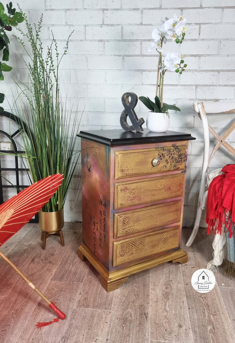 Moonshine metallic is the perfect paint to add shimmer and shine to your chalk painted furniture. Pink, blue and gold metal moonshine metallic painted bedside table with Cherry blossom rub on transfer. Staged in a room with plants and decorative black and symbol.
