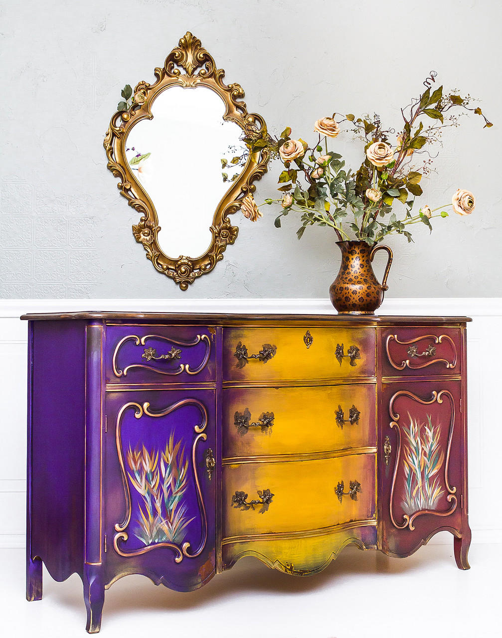 Dixie Belle Paint Muscadine Wine Chalk Mineral Paint is the perfect paint for any DIY project! Purple yellow and dark red chalk painted dresser with wheat style plants painted on. Staged with mirror and cheetah printed vase with roses.