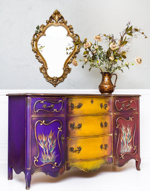 Dixie Belle Paint Muscadine Wine Chalk Mineral Paint is the perfect paint for any DIY project! Purple yellow and dark red chalk painted dresser with wheat style plants painted on. Staged with mirror and cheetah printed vase with roses.