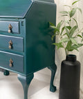 Dixie Belle Paint Palmetto Chalk Mineral Paint is the perfect paint for any DIY project! Up-close side view of a Green chalk painted secretary desk with gold hardware. Staged next to a black vase with a plant inside.