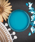 Dixie Belle Paint Peacock Chalk Mineral Paint is the perfect paint for any DIY project! Top view of open blue chalk mineral paint. Staged next to flowers and blue glass.