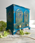 Dixie Belle Paint Peacock Chalk Mineral Paint is the perfect paint for any DIY project! Blue chalk mineral painted jewelry box with gold in the corners and gold hardware, with peacock rub on transfers. Staged next to greenery.