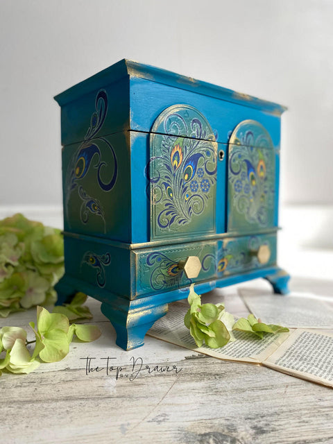 Dixie Belle Paint Peacock Chalk Mineral Paint is the perfect paint for any DIY project! Blue chalk mineral painted jewelry box with gold in the corners and gold hardware, with peacock rub on transfers. Staged next to greenery.