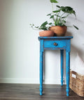 Dixie Belle Paint Peacock Chalk Mineral Paint is the perfect paint for any DIY project! Blue chalk painted end table with red chalk paint blended to make it look distressed. Staged with clay vase and plants and a wicker basket with pillow.