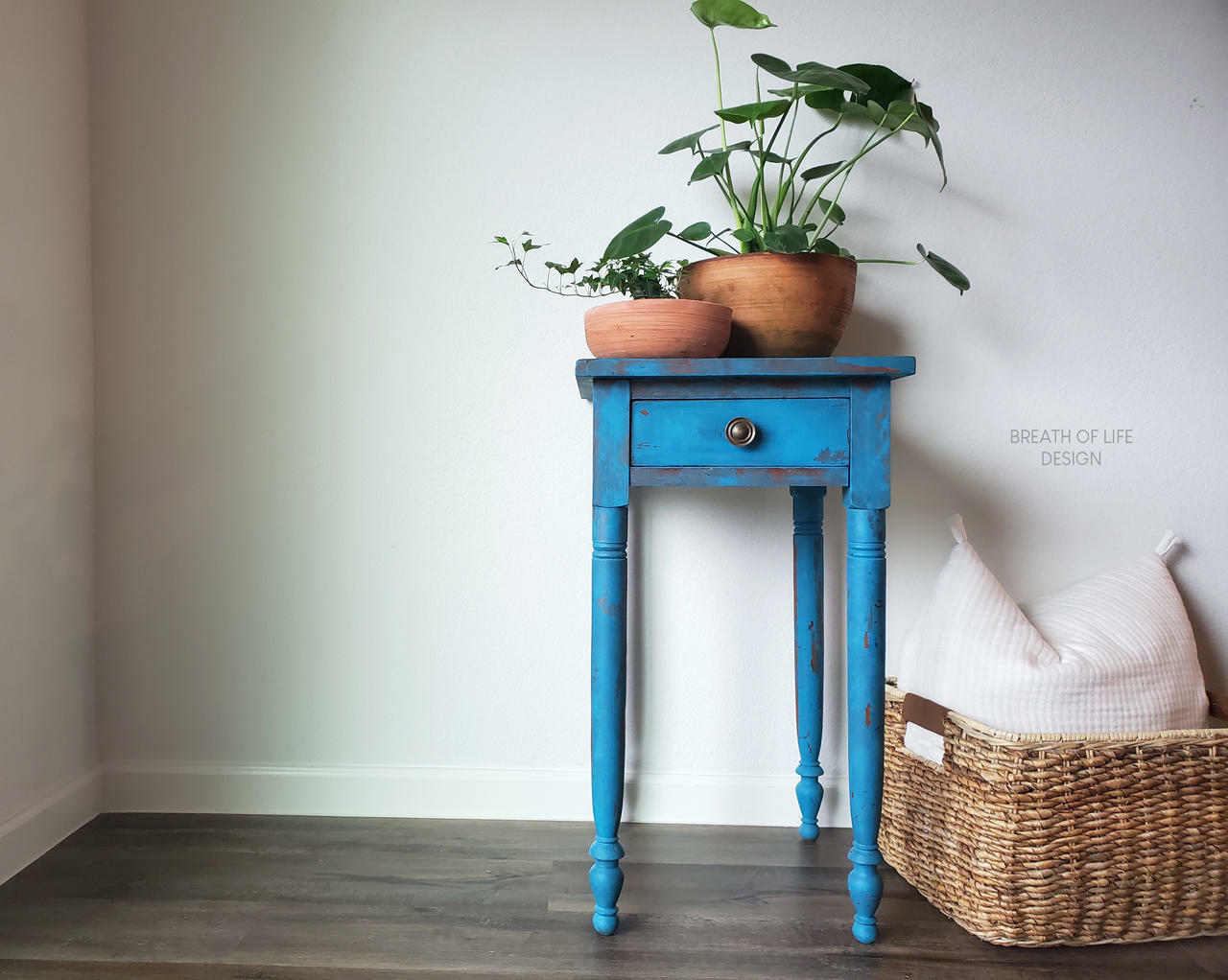 Dixie Belle Paint Peacock Chalk Mineral Paint is the perfect paint for any DIY project! Blue chalk painted end table with red chalk paint blended to make it look distressed. Staged with clay vase and plants and a wicker basket with pillow.