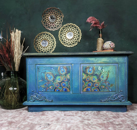 Dixie Belle Paint Peacock Chalk Mineral Paint is the perfect paint for any DIY project! Blue chalk painted blanket box with peacock rub on transfers and scroll wood applique. Staged with glass vase and plants inside.