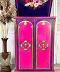 Dixie Belle Paint Peony Chalk Mineral Paint is the perfect paint for any DIY project! Pink chalk painted open top cabinet blended with purple around the doors in front with gold on the trim and a floral rub on transfer on the top of the cabinet door. Staged next to faux wheat plants in a vase.
