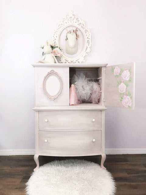 Dixie Belle Paint Pink Champagne Chalk Mineral Paint is the perfect paint for any DIY project! Light pink chalk painted armoire with a white blend and roses painted on the inside of the doors. Staged with little girl clothes and toys.