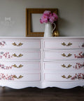 Dixie Belle Paint Pink Champagne Chalk Mineral Paint is the perfect paint for any DIY project! Light pink chalk painted dresser with floral rub on transfers on the drawers and gold hardware. Staged with pink flowers in a white vase.