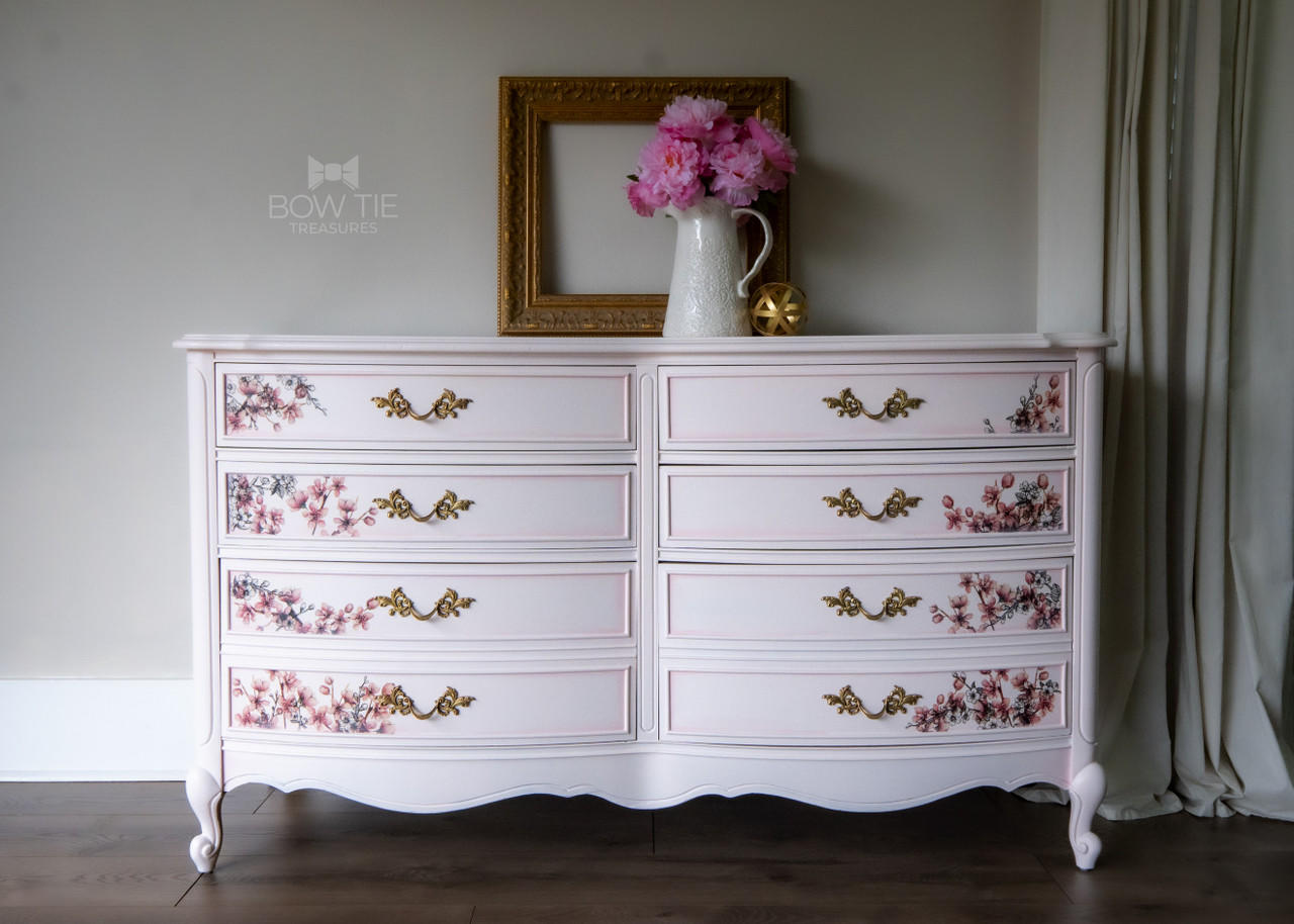 Dixie Belle Paint Pink Champagne Chalk Mineral Paint is the perfect paint for any DIY project! Light pink chalk painted dresser with floral rub on transfers on the drawers and gold hardware. Staged with pink flowers in a white vase.