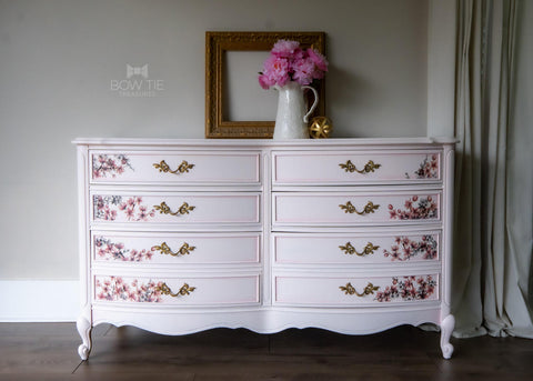 Dixie Belle Paint Pink Champagne Chalk Mineral Paint is the perfect paint for any DIY project! Light pink chalk painted dresser with floral rub on transfers on the drawers and gold hardware. Staged with pink flowers in a white vase.